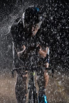 A triathlete braving the rain as he cycles through the night, preparing himself for the upcoming marathon. The blurred raindrops in the foreground and the dark, moody atmosphere in the background add to the sense of determination and grit shown by the athlete