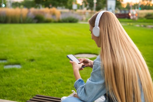 Blonde girl sits with her back in headphones and uses a mobile phone in a clearing in the park.