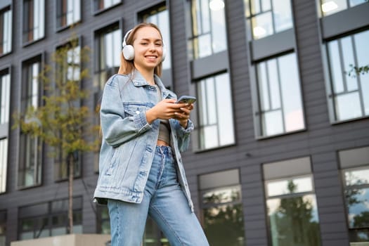 Young blonde woman in denim in headphones listens to music in the street.