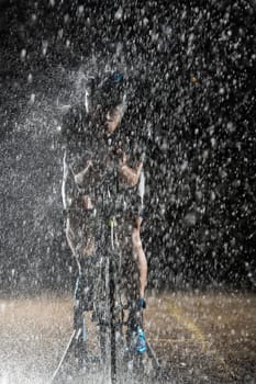 A triathlete braving the rain as he cycles through the night, preparing himself for the upcoming marathon. The blurred raindrops in the foreground and the dark, moody atmosphere in the background add to the sense of determination and grit shown by the athlete