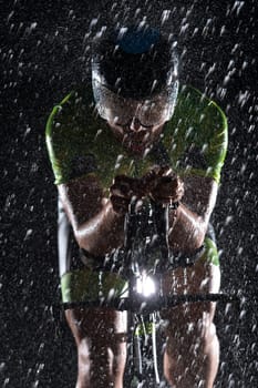 A triathlete braving the rain as he cycles through the night, preparing himself for the upcoming marathon. The blurred raindrops in the foreground and the dark, moody atmosphere in the background add to the sense of determination and grit shown by the athlete