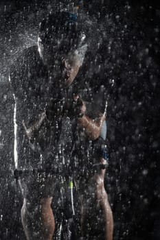 A triathlete braving the rain as he cycles through the night, preparing himself for the upcoming marathon. The blurred raindrops in the foreground and the dark, moody atmosphere in the background add to the sense of determination and grit shown by the athlete