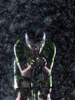 A triathlete braving the rain as he cycles through the night, preparing himself for the upcoming marathon. The blurred raindrops in the foreground and the dark, moody atmosphere in the background add to the sense of determination and grit shown by the athlete