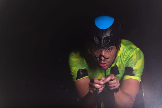 A triathlete braving the rain as he cycles through the night, preparing himself for the upcoming marathon. The blurred raindrops in the foreground and the dark, moody atmosphere in the background add to the sense of determination and grit shown by the athlete