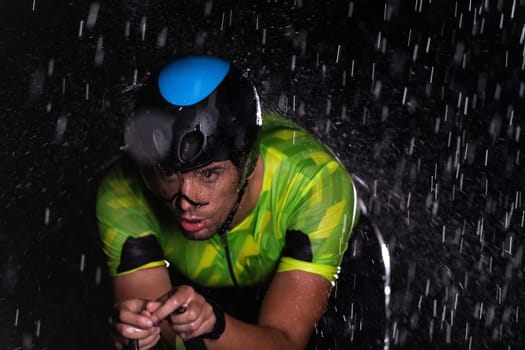 A triathlete braving the rain as he cycles through the night, preparing himself for the upcoming marathon. The blurred raindrops in the foreground and the dark, moody atmosphere in the background add to the sense of determination and grit shown by the athlete