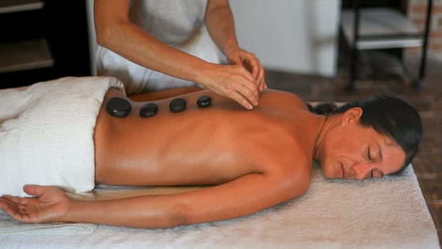 professional caucasian female physiotherapist masseuse performing stone massage on backbone to hispanic latino brunette woman in spa clinic