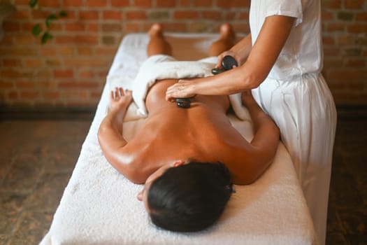 professional caucasian female physiotherapist masseuse performing stone massage on backbone to hispanic latino brunette woman in spa clinic