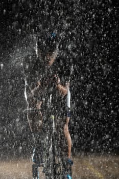 A triathlete braving the rain as he cycles through the night, preparing himself for the upcoming marathon. The blurred raindrops in the foreground and the dark, moody atmosphere in the background add to the sense of determination and grit shown by the athlete