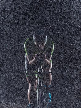 A triathlete braving the rain as he cycles through the night, preparing himself for the upcoming marathon. The blurred raindrops in the foreground and the dark, moody atmosphere in the background add to the sense of determination and grit shown by the athlete