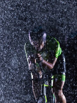 A triathlete braving the rain as he cycles through the night, preparing himself for the upcoming marathon. The blurred raindrops in the foreground and the dark, moody atmosphere in the background add to the sense of determination and grit shown by the athlete