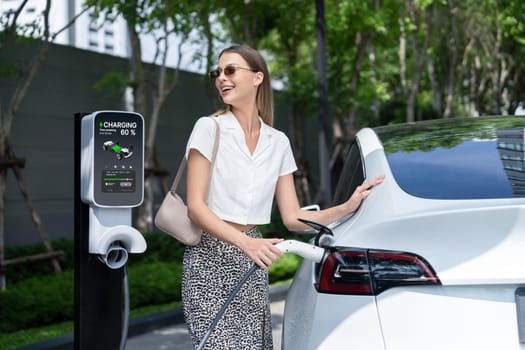 Young woman travel with EV electric car charging in green sustainable city outdoor garden in summer. Urban sustainability lifestyle by green clean rechargeable energy of electric BEV vehicle innards