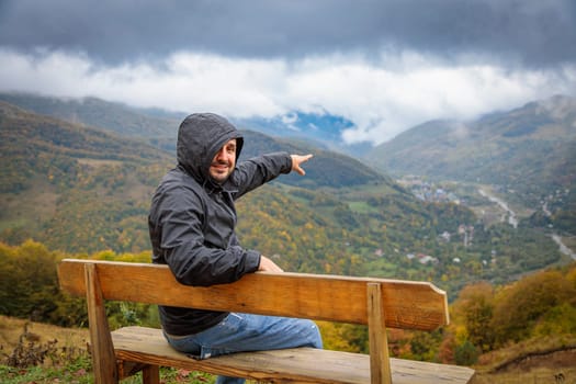 A man is sitting on a wooden bench on an observation deck, overlooking a stunning mountain range. He is peacefully enjoying the serene natural beauty and taking in the scenic views.