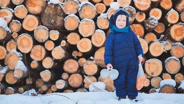 A boy stands by the logs in winter with mugs and tangerines