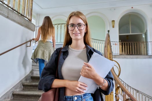 Portrait of young teenage girl student posing inside an educational building, standing on stairs. Smiling female with bag, textbook looking at camera. Youth, education, college, university