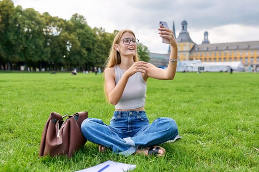 Teenage girl student having video call on smartphone, resting sitting on grass lawn near university building. Technology, leisure, communication, youth