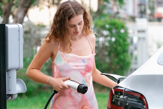 Modern eco-friendly woman portrait recharging electric vehicle from home EV charging station. EV car technology utilized for home resident to future environmental sustainability. Synchronos