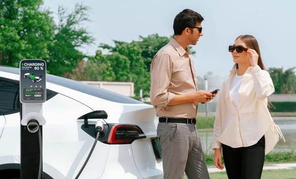 Young couple use smartphone to pay for electricity at public EV car charging station green city park. Modern environmental and sustainable urban lifestyle with EV vehicle. Expedient