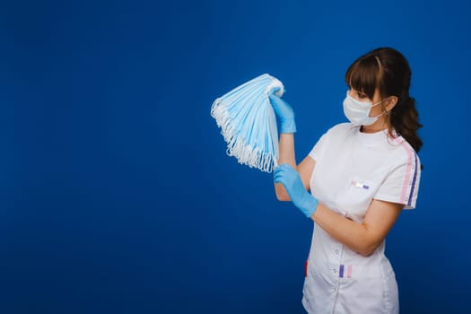 A young girl doctor in a medical mask holds a lot of masks in her hands. A nurse with white masks in her hands on a blue background.