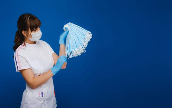 a doctor girl in a medical mask on a blue background holds a lot of protective masks in her hands.