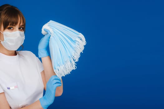 A young girl doctor in a medical mask holds a lot of masks in her hands. A nurse poses on a blue background.