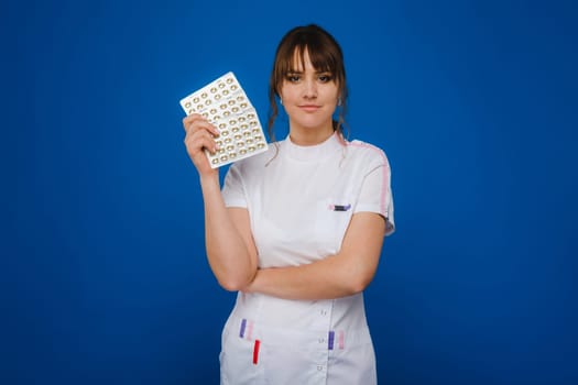 The concept of health care. A young brunette doctor in a white coat on a blue background shows plates with capsules to take
