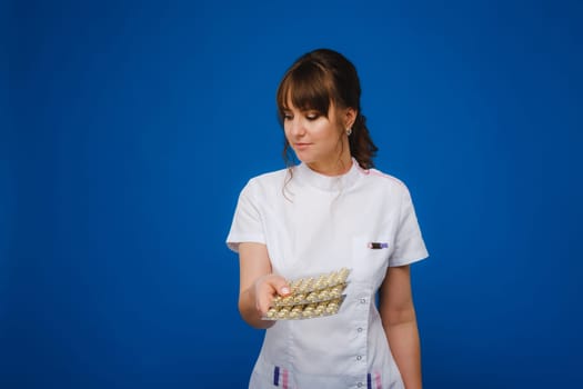 The concept of health care. A young brunette doctor in a white coat on a blue background shows plates with capsules to take