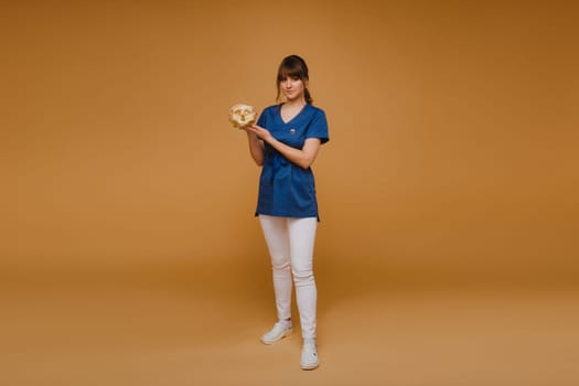 Cute young doctor girl holding a human skull, brown background behind the doctor.