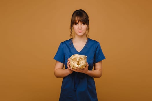 Cute young doctor girl holding a human skull, brown background behind the doctor.