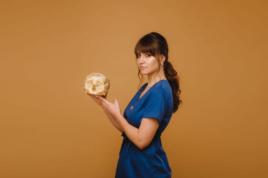 Cute young doctor girl holding a human skull, brown background behind the doctor.