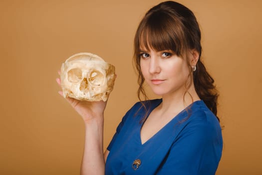 Cute young doctor girl holding a human skull, brown background behind the doctor.