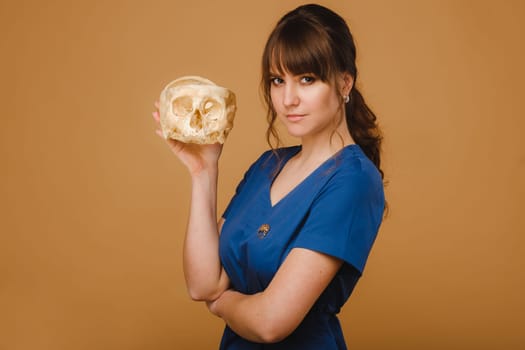Cute young doctor girl holding a human skull, brown background behind the doctor.