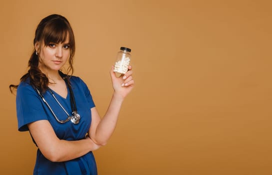 A doctor in a blue lab coat holds a jar of capsules in his hands on a brown background.