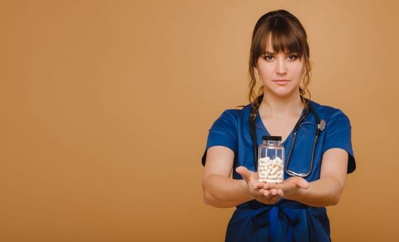 A doctor in a blue lab coat holds a jar of capsules in his hands on a brown background.