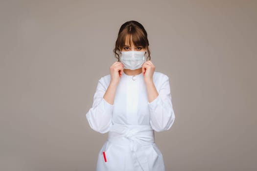 A girl doctor stands in a medical mask, isolated on a gray background