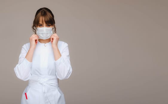 A girl doctor stands in a medical mask on a gray background.