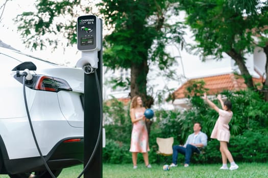 Focus electric vehicle recharge from home charging station on blur background of happy and playful family playing together. EV car using alternative and sustainable energy for better future.Synchronos