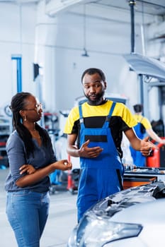 Qualified mechanic helping client with car maintenance in auto repair shop. Employee in garage facility looking over automobile parts with woman, mending her vehicle radiator during inspection