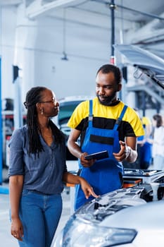 Mechanic in car service uses tablet to calculate invoice after fixing broken automobile motor. Certified auto repair shop worker uses device to inform woman of final costs after repairing vehicle
