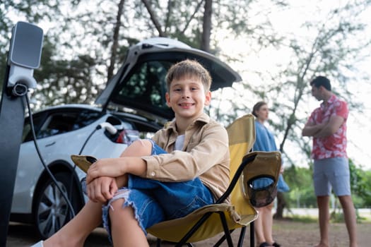 Little boy portrait sitting on camping chair with his family in background. Road trip travel with alternative energy charging station for eco-friendly car concept. Perpetual