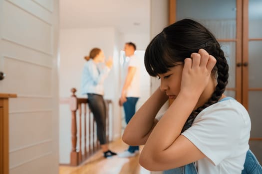 Stressed and unhappy young girl huddle in corner, cover her ears blocking sound of her parent arguing in background. Domestic violence at home and traumatic childhood develop to depression. Synchronos