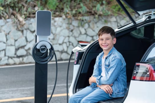 Little boy sitting on car trunk while recharging eco-friendly electric car from EV charging station. EV car road trip travel concept for alternative transportation powered sustainable energy.Perpetual