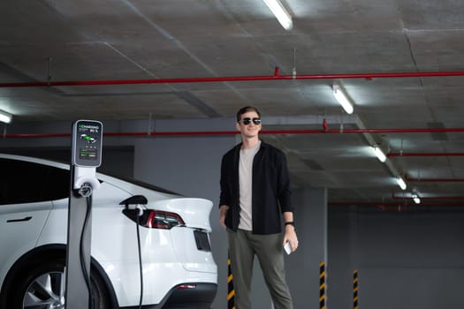 Young man travel with EV electric car to shopping center parking lot charging in downtown city showing urban sustainability lifestyle by green clean rechargeable energy of electric vehicle innards