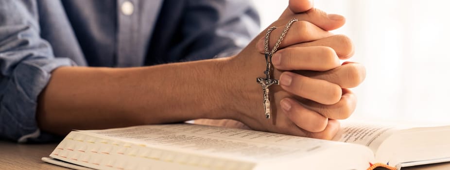 Asian male folded hand prayed on holy bible book while holding up a pendant crucifix. Spiritual, religion, faith, worship, christian and blessing of god concept. Blurring background. Burgeoning.