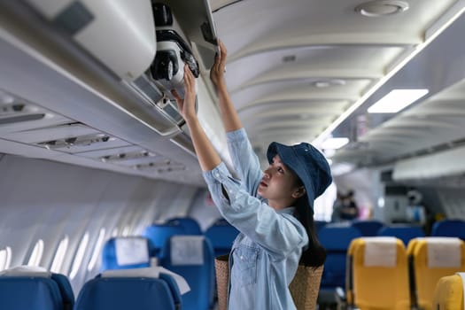 Young woman putting luggage into overhead locker on airplane. Traveler placing carry on bag in overhead compartment in aircraft.