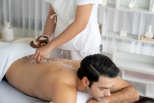 Blissful man customer having exfoliation treatment in luxury spa salon with warmth candle light ambient. Salt scrub beauty treatment in health spa body scrub. Quiescent