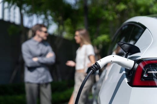 Young couple travel with EV electric car charging in green sustainable city outdoor garden in summer shows urban sustainability lifestyle by green clean rechargeable energy of electric vehicle innards