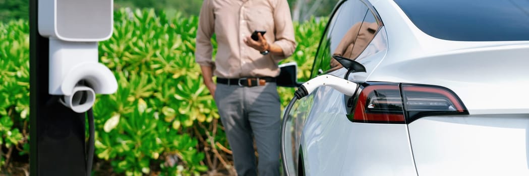 Young man use smartphone to pay for electricity at public EV car charging station green city park. Modern environmental and sustainable urban lifestyle with EV vehicle. Panorama Expedient