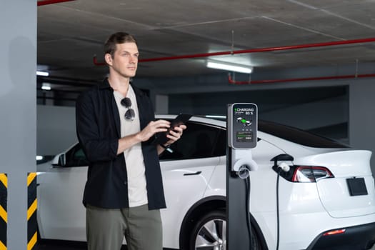 Young man travel with EV electric car to shopping center parking lot charging in downtown city showing urban sustainability lifestyle by green clean rechargeable energy of electric vehicle innards