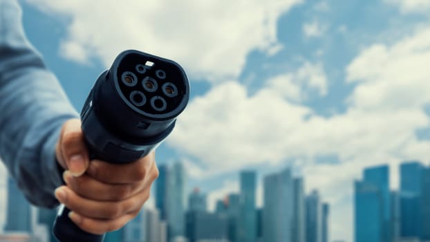 Focus EV charger pointing in front of camera with businessman's hand in blurry background of cityscape and cloud. Electric car charger plug and alternative clean energy reducing CO2 emission.Peruse