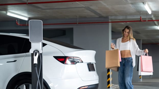 Young woman travel with EV electric car to shopping center parking lot charging in downtown city showing urban sustainability lifestyle by green clean rechargeable energy of electric vehicle innards